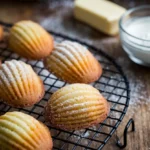 Freshly baked golden madeleine cookies on a cooling rack, lightly dusted with powdered sugar.